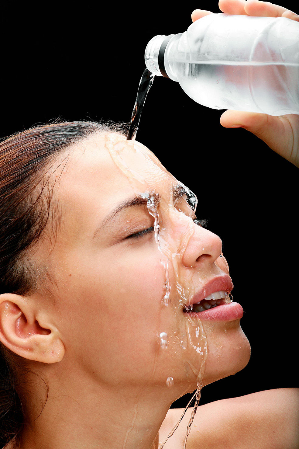 Esta imagen captura un instante de frescura y vitalidad en un primer plano impactante. Una mujer con la piel radiante vierte agua sobre su rostro con los ojos cerrados, disfrutando de una sensación de hidratación y bienestar. Las gotas resbalan suavemente por su piel, resaltando su textura natural y la pureza del momento.  El fondo negro crea un poderoso contraste, haciendo que la luminosidad de su piel y la transparencia del agua cobren protagonismo. Su expresión relajada y su leve sonrisa transmiten una sensación de frescura, energía y revitalización, evocando la importancia de la hidratación y el cuidado de la piel.  Esta imagen es una representación perfecta de la sensación refrescante que aportan los tratamientos de limpieza profunda e hidratación, resaltando la belleza natural en su estado más puro.