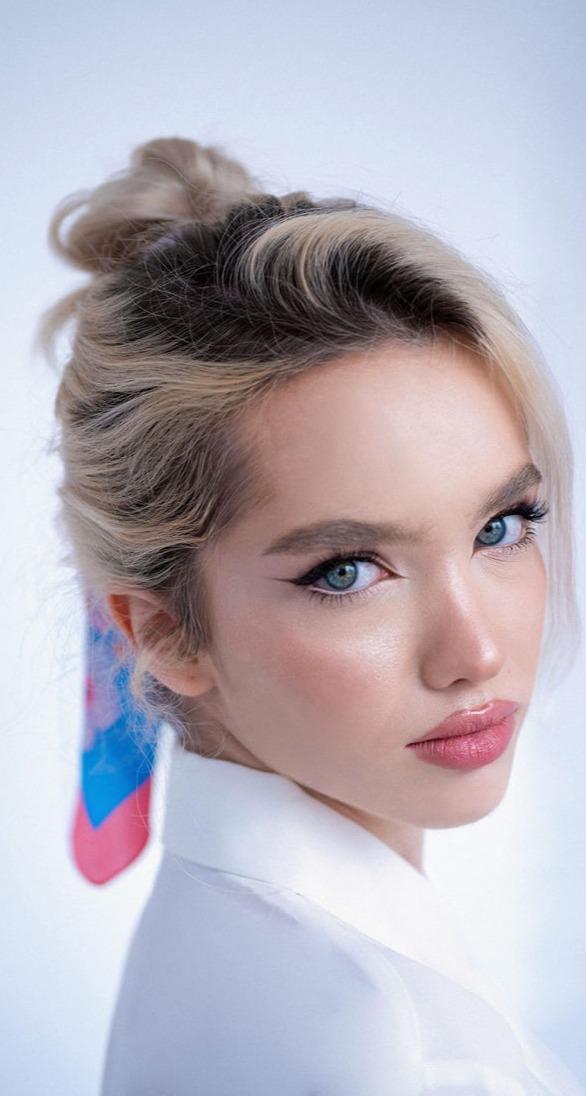 Esta imagen presenta un retrato sofisticado y moderno de una mujer con una mirada impactante y una expresión llena de confianza. Sus ojos azul intenso están enmarcados por un delineado felino que acentúa su profundidad y magnetismo. Su piel luce impecable, con un brillo natural que resalta su estructura ósea y aporta frescura al conjunto.  Su cabello, recogido en un moño desenfadado, deja ver mechones sueltos que aportan dinamismo a la imagen. Un pañuelo de colores vibrantes atado en su cabello añade un toque de estilo y sofisticación. Sus labios, con un acabado jugoso en un tono rosado, equilibran la intensidad de su mirada y refuerzan la armonía de su maquillaje.  El fondo claro y minimalista resalta aún más su presencia, mientras que la iluminación suave y difusa aporta una sensación etérea y elegante. La composición transmite una belleza moderna, refinada y empoderada.