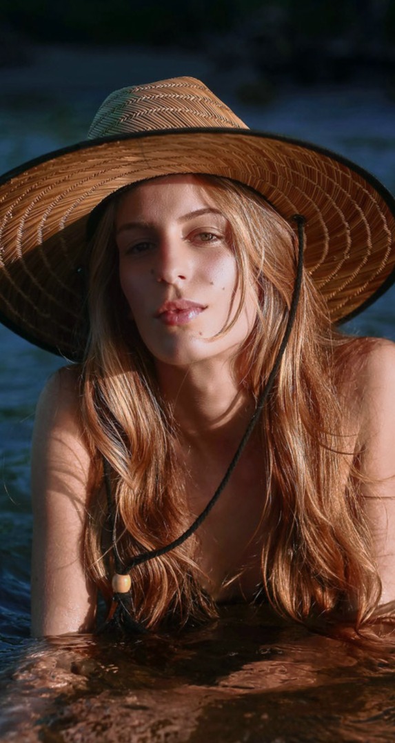 Esta imagen captura la esencia de la belleza natural y la serenidad en un entorno acuático. Una mujer con cabello dorado y suelto se encuentra sumergida en el agua hasta los hombros, con un gran sombrero de paja que la protege del sol. Su expresión relajada y su mirada directa transmiten confianza y tranquilidad, mientras los reflejos dorados del atardecer resaltan la calidez de su piel y la textura de su cabello.  El contraste entre la luz y la sombra en su rostro aporta profundidad a la imagen, mientras que el fondo de agua en tonos azulados refuerza la sensación de frescura y conexión con la naturaleza. El detalle del cordón del sombrero, que cae con naturalidad sobre su pecho, añade un toque bohemio y aventurero a la escena.  Esta fotografía evoca el equilibrio perfecto entre el bienestar, la libertad y el disfrute del sol y el agua, reflejando un momento de paz y armonía con el entorno.