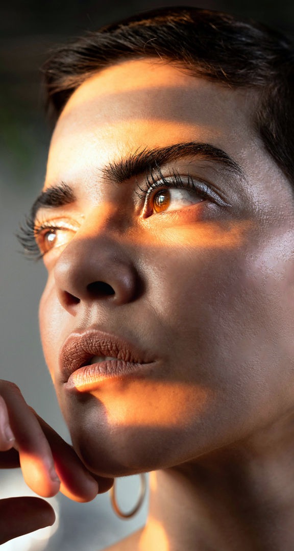Esta imagen transmite una intensidad única a través del juego de luces y sombras sobre el rostro. Una mujer de mirada profunda es iluminada por los cálidos rayos del sol, que se filtran en franjas a través de una ventana, resaltando la textura natural de su piel y la expresividad de sus ojos marrón ámbar.  Su piel luce radiante y auténtica, con un brillo sutil que enfatiza su belleza natural. Sus cejas definidas y pestañas largas enmarcan su mirada, mientras sus labios en tono tierra agregan armonía y calidez a la composición.  La postura con la mano suavemente apoyada cerca del rostro refuerza la sensación de introspección y fortaleza. La imagen captura un momento de reflexión, transmitiendo confianza, serenidad y una conexión con la luz y el entorno.  Esta fotografía evoca el poder de la belleza natural, resaltando la elegancia de la simplicidad y la autenticidad en cada detalle.
