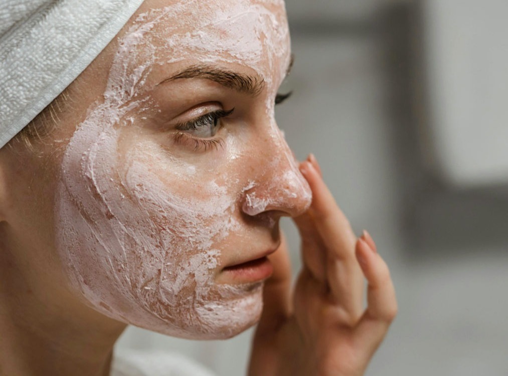 La imagen muestra a una mujer aplicándose una mascarilla facial cremosa de color blanco con tonos rosados. Lleva una toalla blanca enrollada en su cabeza, lo que indica que está en una rutina de cuidado de la piel, posiblemente después de una ducha o baño. Su expresión es relajada y concentrada mientras se toca suavemente el rostro, asegurándose de distribuir bien el producto. La iluminación tenue y el fondo desenfocado sugieren un ambiente de spa o de relajación en casa. La textura de la mascarilla es visible, mostrando su efecto hidratante o exfoliante sobre la piel.