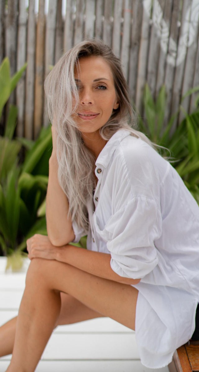 Esta imagen captura un retrato natural y sereno de una mujer con una belleza relajada y auténtica. Su piel luce fresca y luminosa, con un aspecto saludable y sin artificios. Su cabello ondulado en tonos dorados cae suavemente sobre sus hombros, enmarcando su rostro con un aire desenfadado pero elegante.  Sostiene una taza blanca en sus manos, evocando una atmósfera de calma y bienestar, como si estuviera disfrutando de un momento de tranquilidad en casa. Su expresión, con una leve sonrisa y una mirada directa, transmite confianza y serenidad.  El fondo, ligeramente desenfocado y con tonos cálidos, refuerza la sensación acogedora de la imagen. La iluminación natural y suave realza los matices de su piel y cabello, dando un efecto de frescura y autenticidad.  Esta fotografía representa a la perfección la belleza en su estado más puro, resaltando la importancia del autocuidado, la hidratación y la armonía con uno mismo.